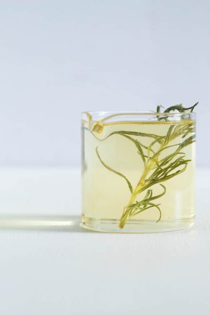 A small jar of pale yellow syrup with a rosemary sprig inside on a white surface.
