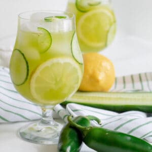 A round glass with green-tinted lemonade and fresh cucumber, jalapeno, and lemon slices as garnish.