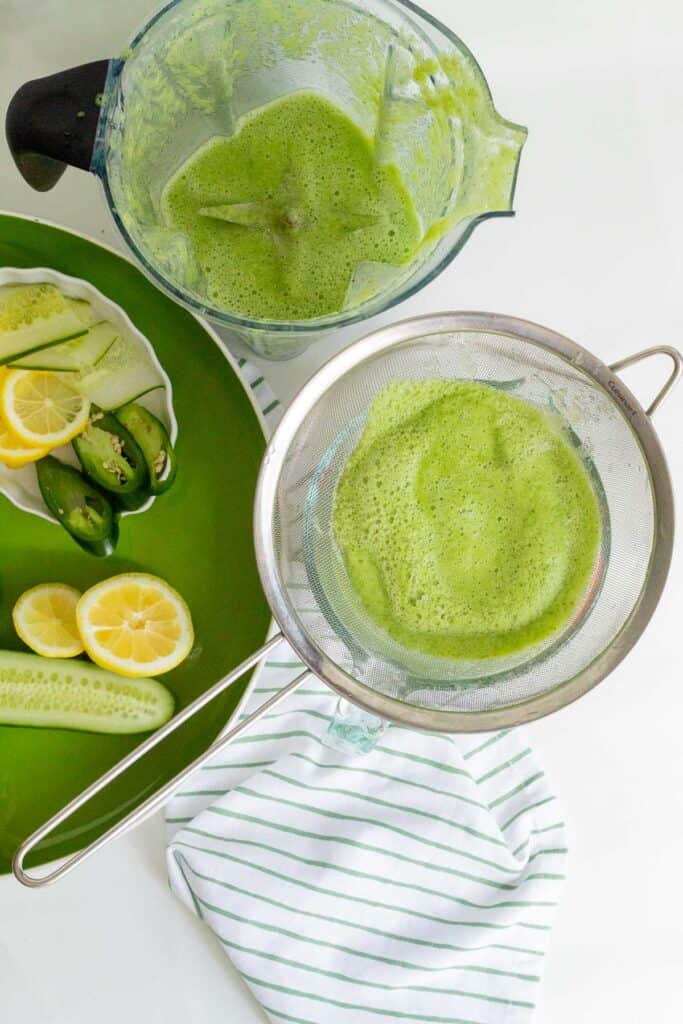Blended cucumber and lemon juice being strained over a pitcher with drink garnishes in the background.