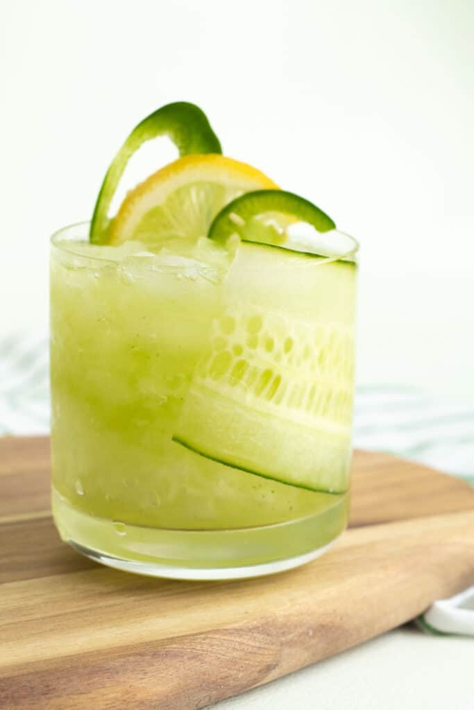 A green cocktail in a small tumbler with fresh cucumber, jalapeno, and lemon slices as garnish.
