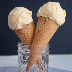 Two ice cream cones with pale yellow scoops of ice cream in front of a blue background.