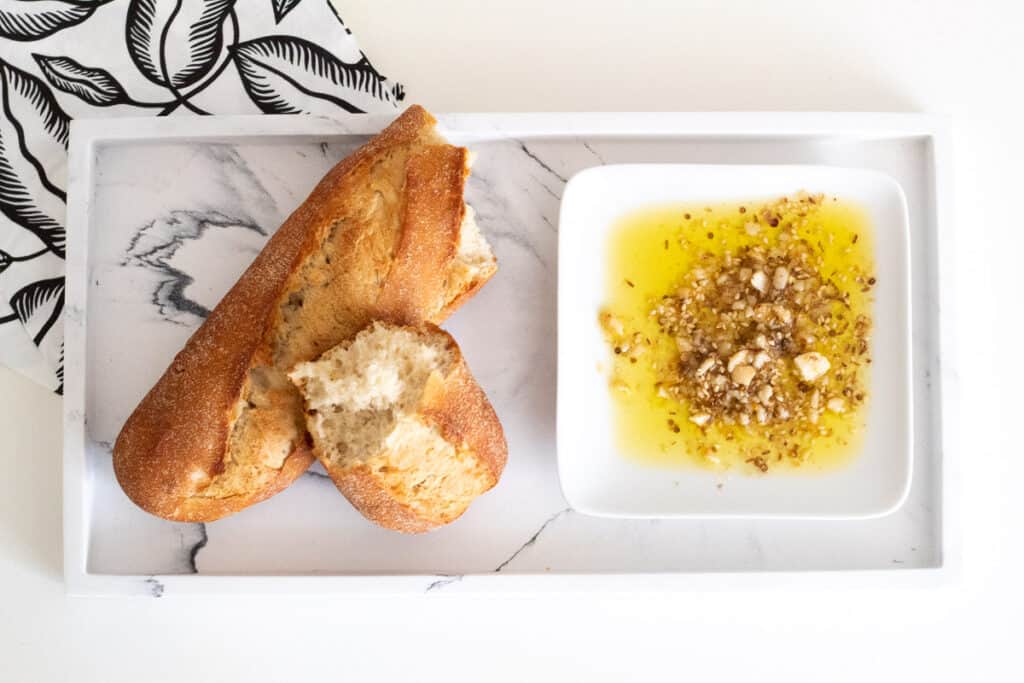 A torn baguette sits next to macadamia dukkah in a dish of olive oil on a marble tray.
