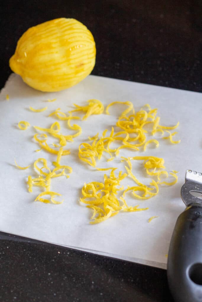 Lemon zest removed from a lemon with a channel zester sits on white parchment paper.