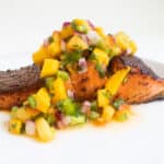 A filet of salmon with a brown crust on a white plate. It is topped with a chunky yellow salsa.