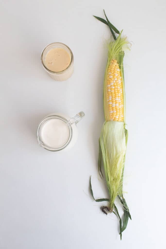 Ingredients for Sweet Corn Ice Cream on a white surface: heavy cream, sweetened condensed milk, and a cob of corn.