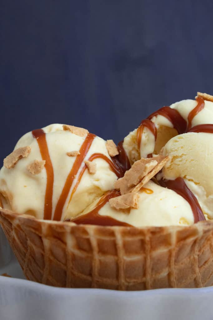 Scoops of pale yellow ice cream are in a waffle cone bowl and drizzled with caramel sauce.