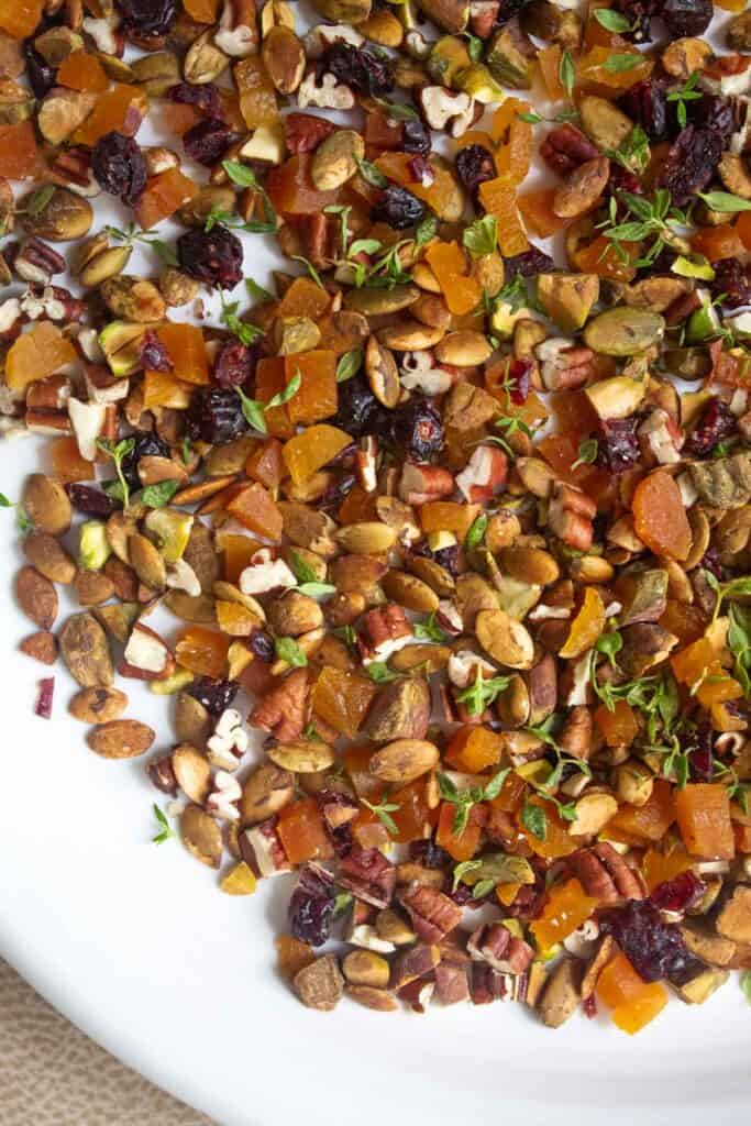 A plate with a mixture of chopped pecans, pistachios, pepitas, apricots, cranberries, and thyme for coating the cheese ball.