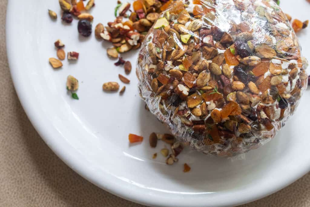 The goat cheese ball coated with chopped pecans, pistachios, apricots, thyme, and cranberries and wrapped in plastic wrap on a white plate.