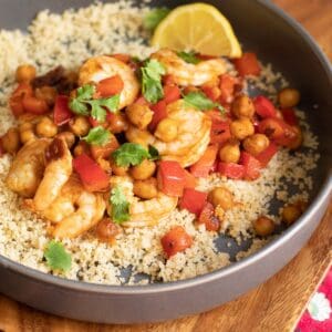 Shrimp, chickpeas, and diced red pepper sitting atop a bed of cous cous garnished with cilantro leaves and lemon.