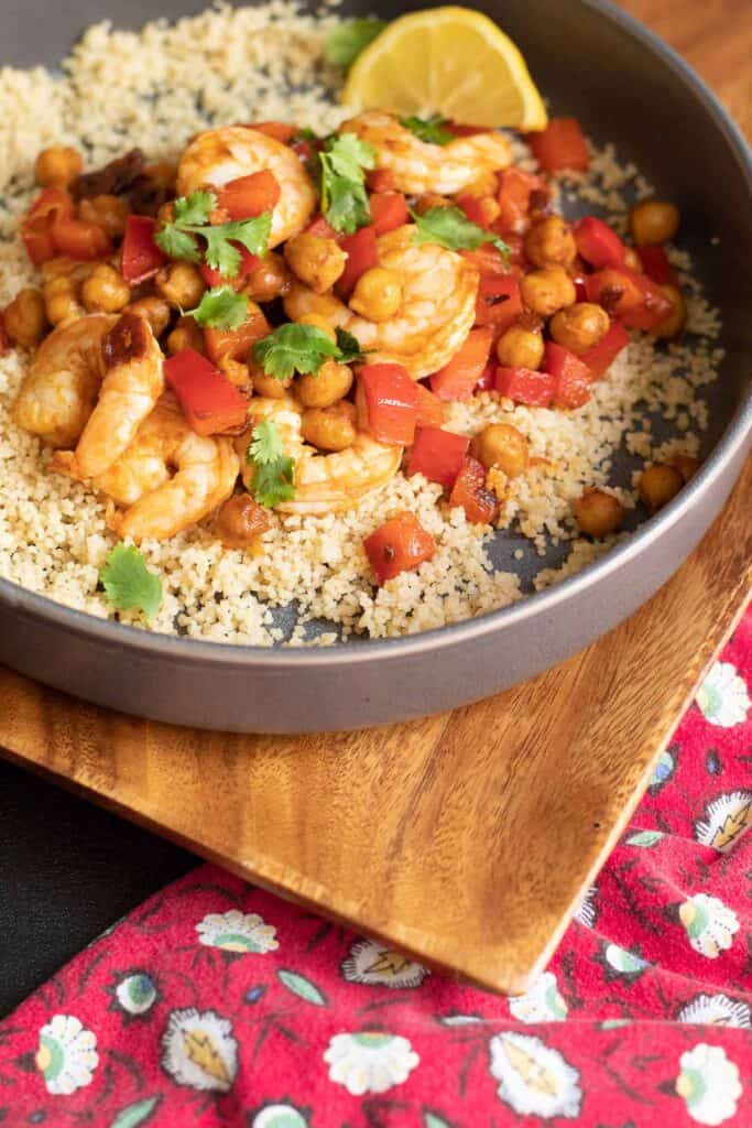 A gray bowl of shrimp, chickpeas, sliced garlic, diced red peppers, and green cilantro over a bed of cous cous sits on a wooden platter with a red floral napkin nearby.