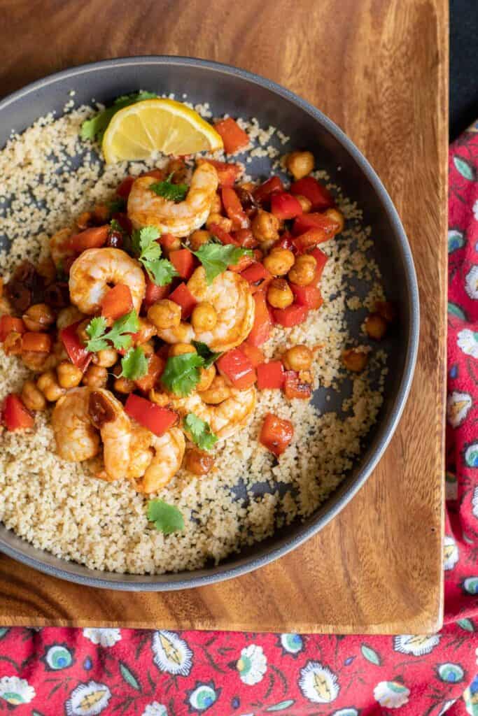 An overhead shot of a gray bowl of shrimp, chickpeas, diced red peppers, and green cilantro over a bed of cous cous sits on a wooden platter with a red floral napkin nearby.