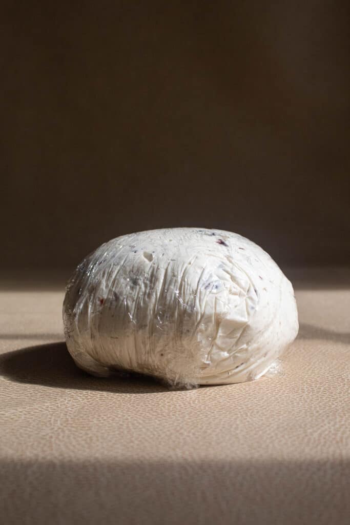 A ball of cream cheese, goat cheese, pepper, and garlic shaped into a ball with plastic wrap on a brown surface.