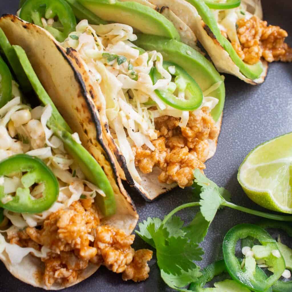 A gray plate with tacos in charred tortillas with pork, avocado, jalapeno, and slaw filling.