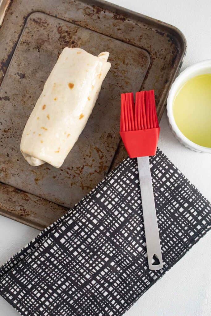 A burrito sits on a baking pan next to a red pastry brush and a small bowl of oil.