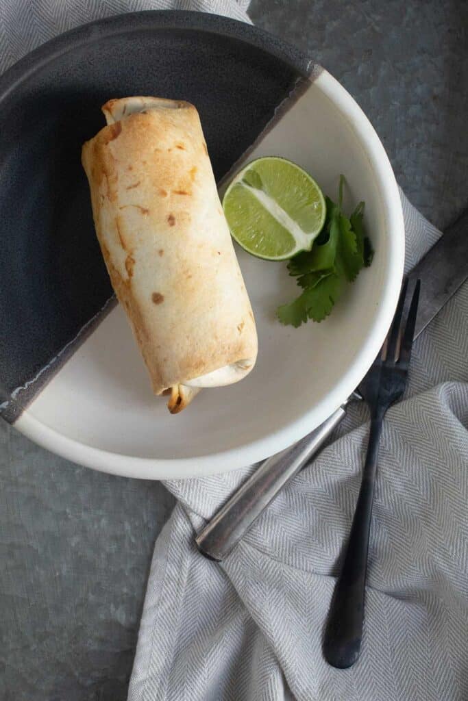 A baked burrito sits in a black and white bowl next to a half of a lime and some leaves of cilantro.