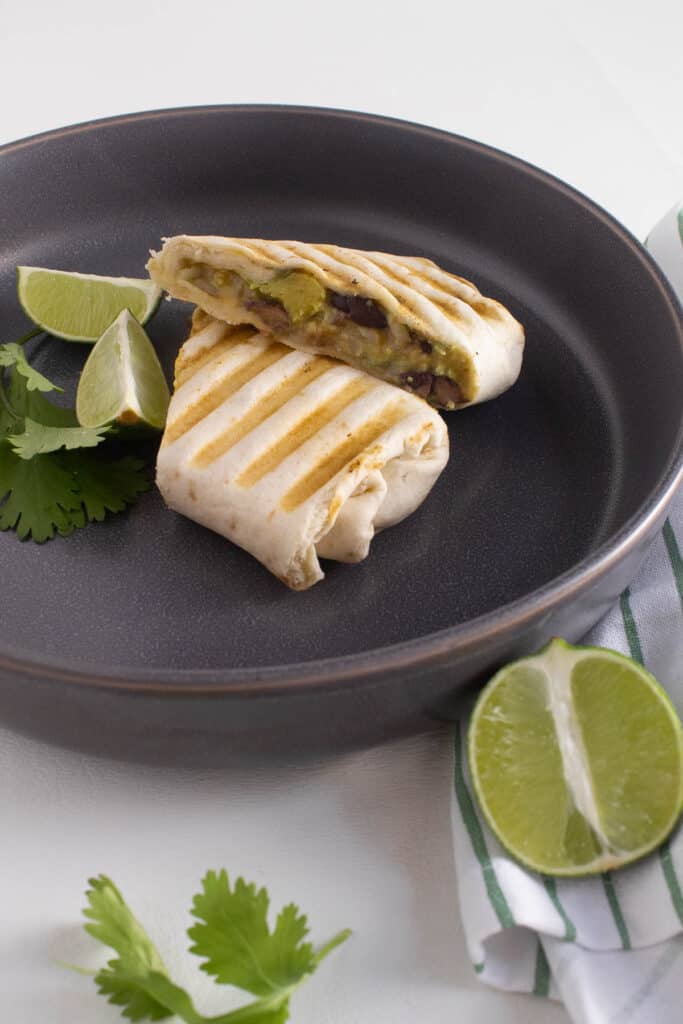 A pressed burrito with grill marks filled with visible chunks of avocado and black beans sits on a gray shallow bowl next to lime wedges and cilantro leaves.