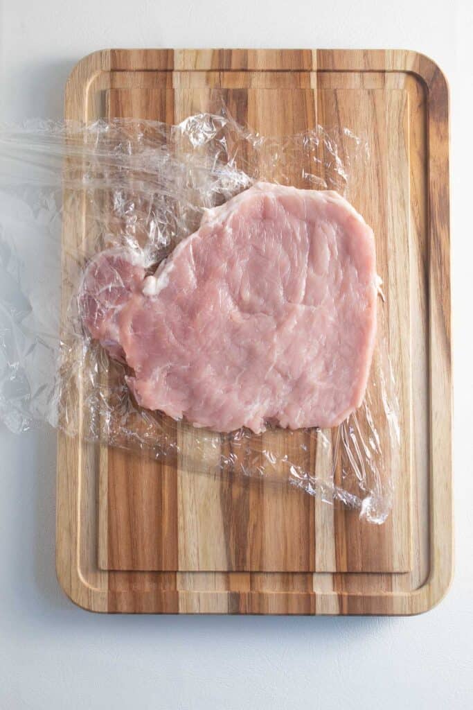 A pork chop that has been pounded thin between two sheets of plastic wrap sits on a wooden cutting board.