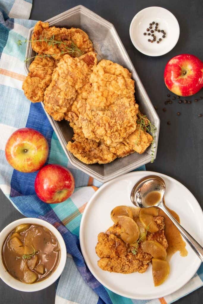 A tray of crispy breaded pork cutlets sits on a dark surface with a bowl of apple cider gravy, apples, peppercorns, and a serving of the pork and gravy nearby.