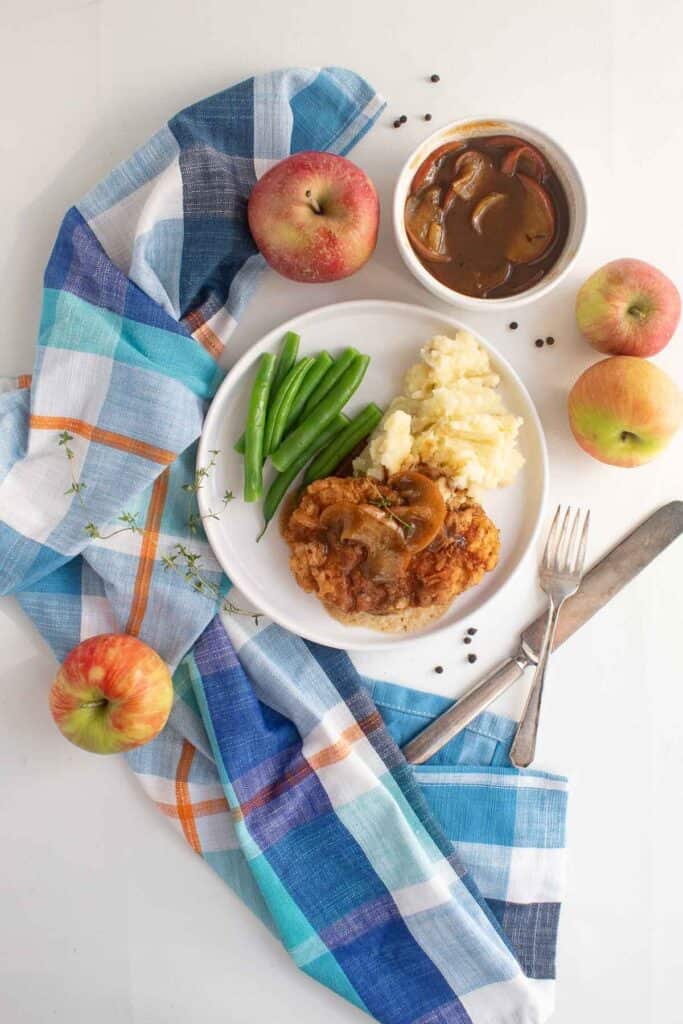 A serving of golden breaded pork and apples on a white plate with green beans and mashed potatoes. A bowl of apple cider gravy and some fresh apples sit alongside the plate.