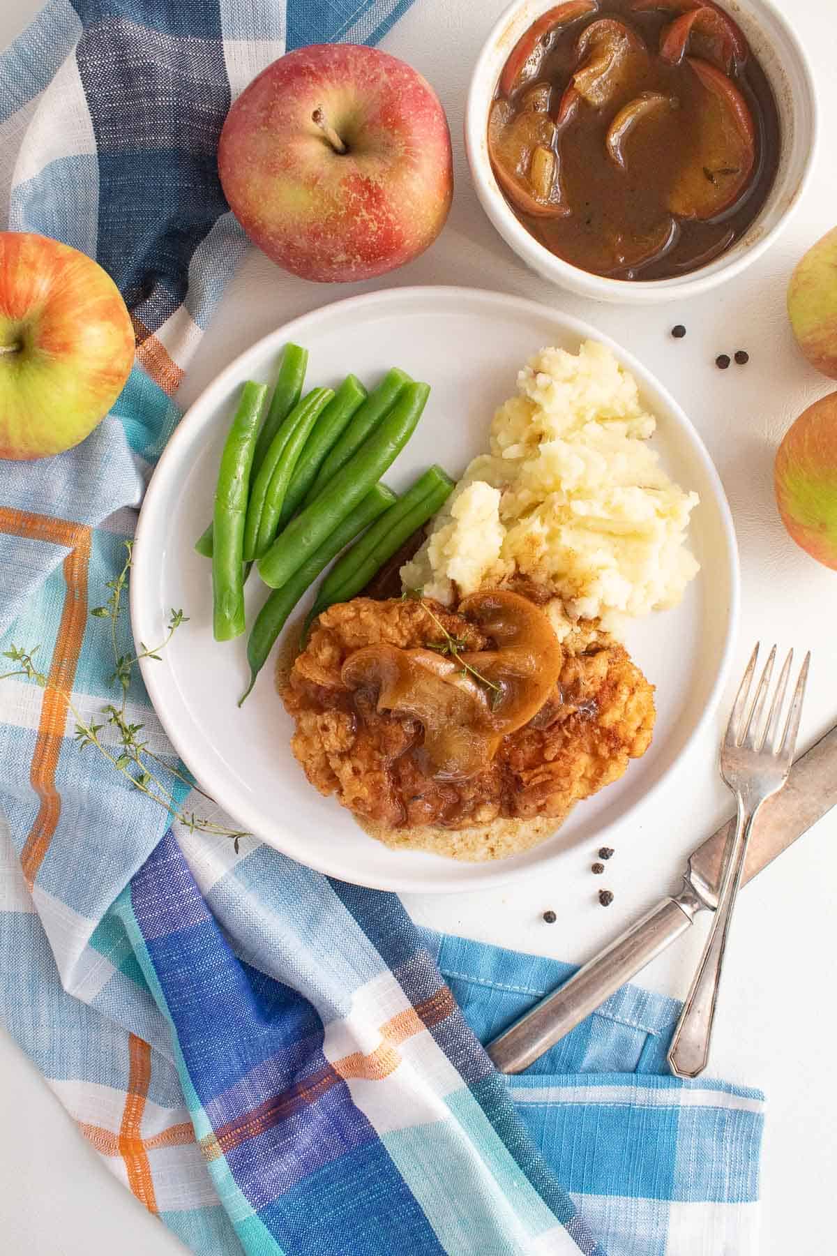 A serving of golden breaded pork and apples on a white plate with green beans and mashed potatoes. A bowl of apple cider gravy and some fresh apples sit alongside the plate.