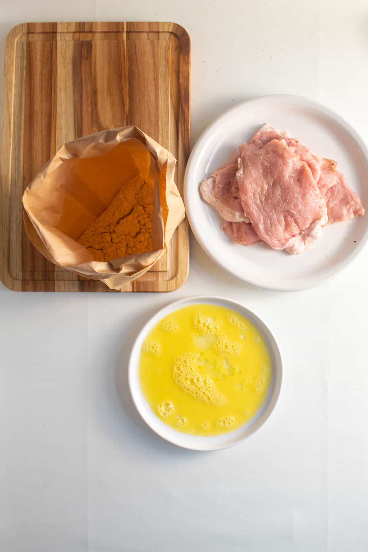 A paper bag of seasoned flour sits on a cutting board alongside a plate of pounded pork chops and a plate of whisked egg for a breading station.