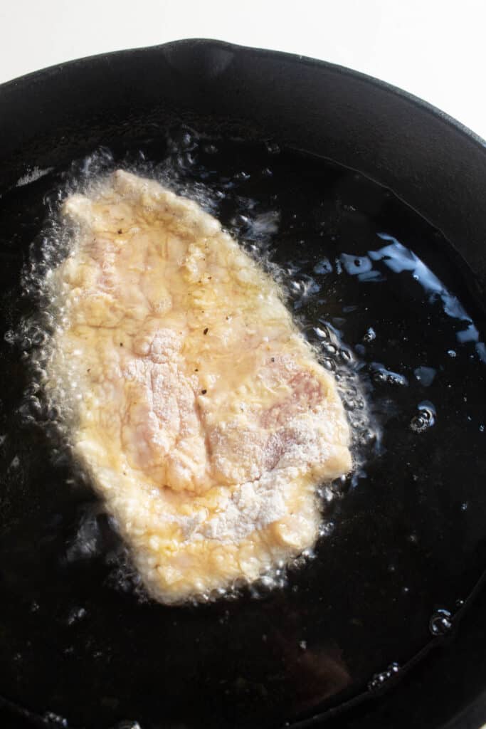 A breaded pork cutlet frying in oil in a cast iron skillet.