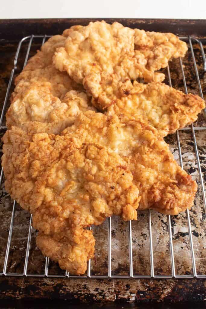 Golden fried pork cutlets sit on a wire rack over a sheet pan.