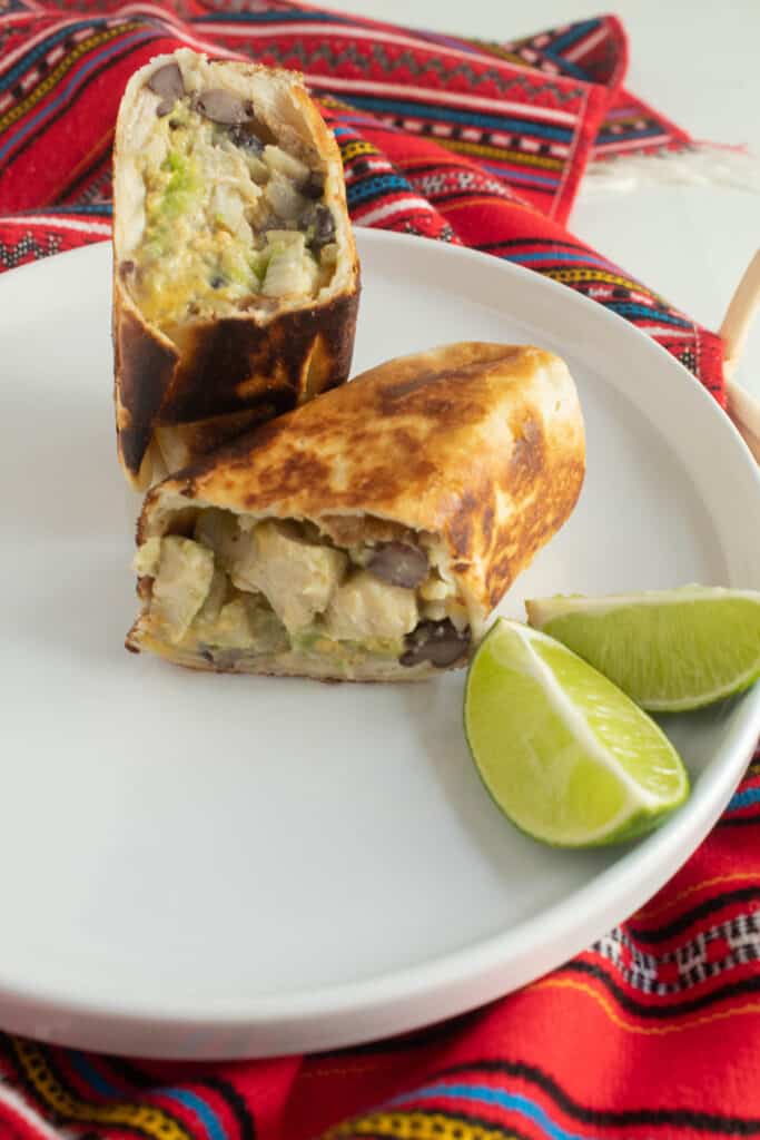 A golden brown tortilla surrounds a filling with chicken chunks, black beans, and avocado. The burrito sits on a white plate next to lime wedges.