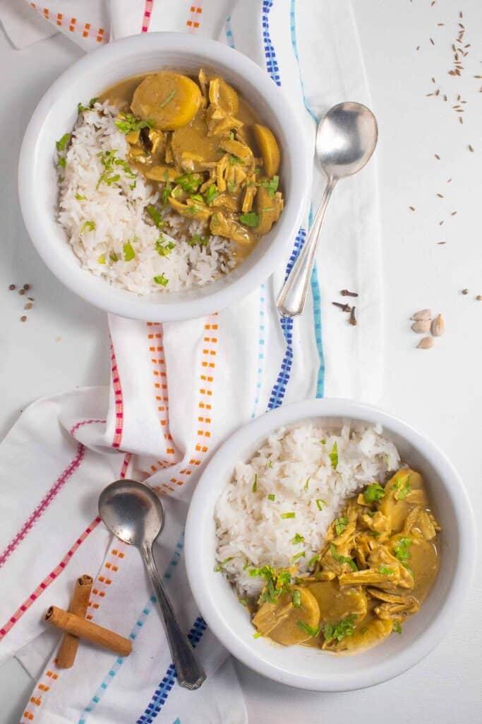 Two bowls of mild coconut curry and rice on a white surface surrounded by whole spices.