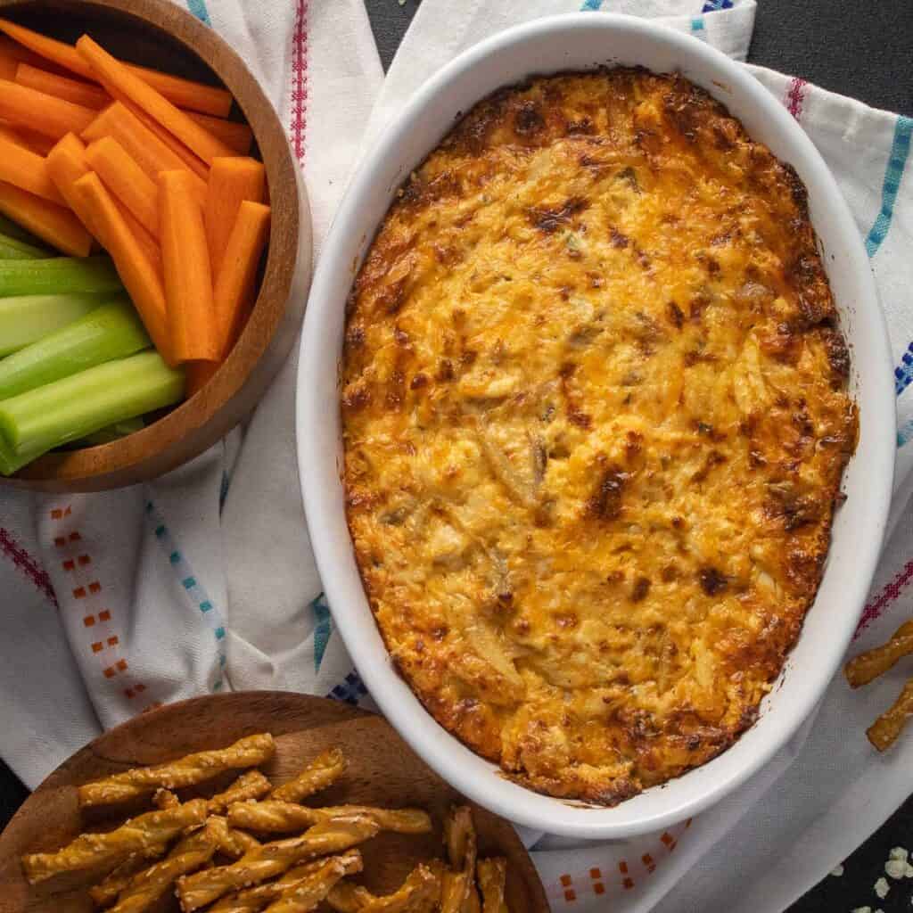 An oval dish of browned cheese dip arranged next to a bowl of carrot and celery sticks and a plate of pretzels.