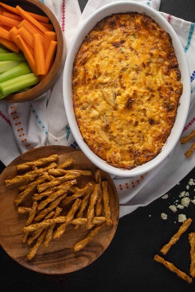 An oval dish of browned cheese dip arranged next to a bowl of carrot and celery sticks, a plate of pretzel sticks, and blue cheese crumbles.