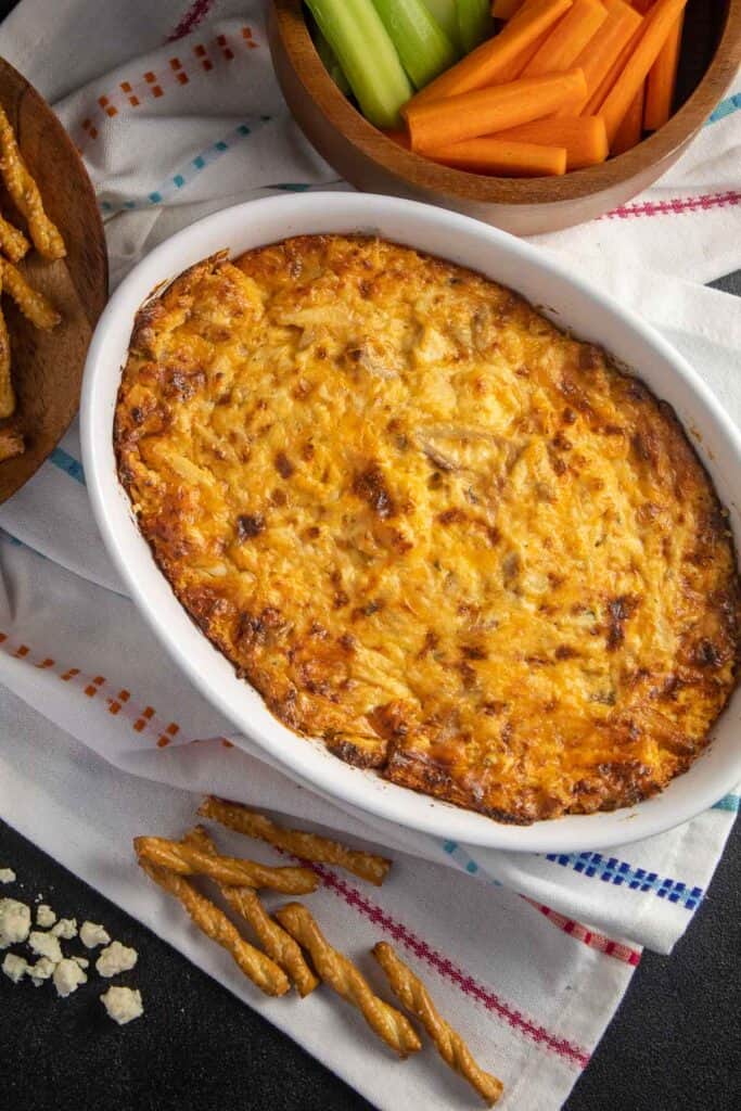 An oval dish of browned cheese dip arranged next to a bowl of carrot and celery sticks, pretzels, and blue cheese crumbles.