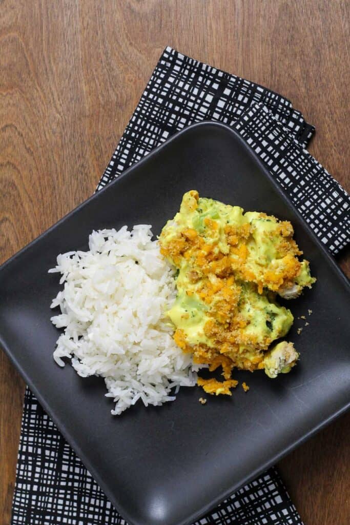 A serving of baked broccoli and chicken divan with curry and a scoop of white rice on a black plate.