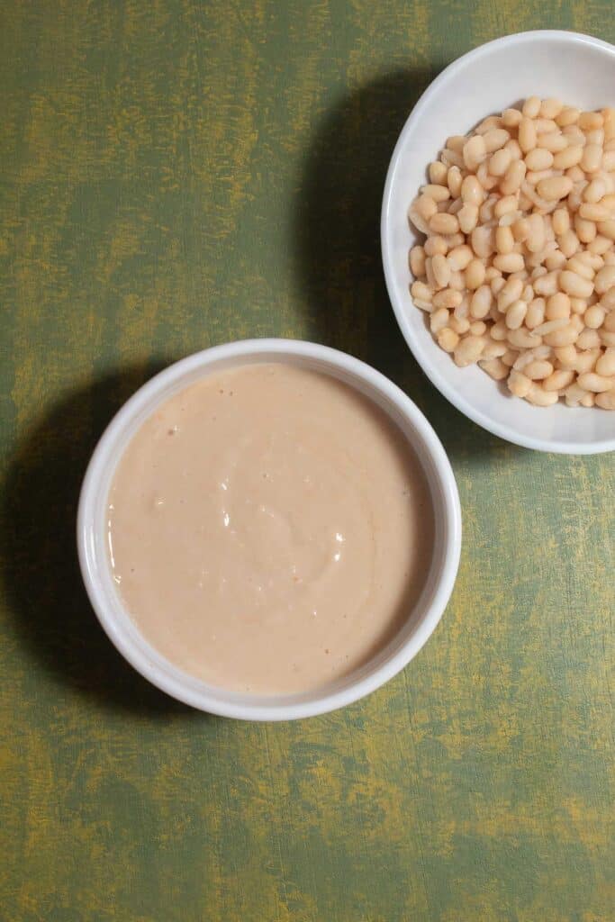 A white bowl containing smoothly pureed beans alongside a bowl of drained and rinsed beans.