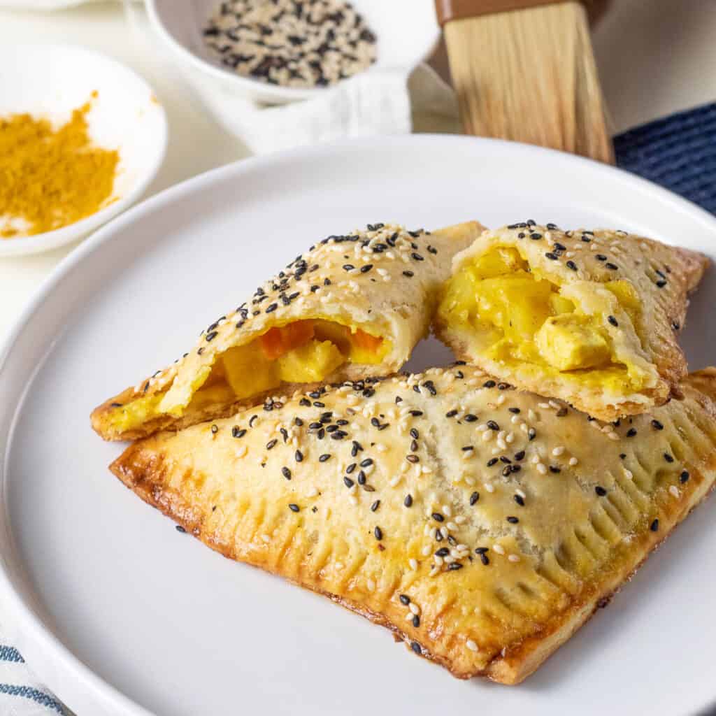 A flaky golden brown sesame seed-flecked turnover with yellow filling rests on a white plate.