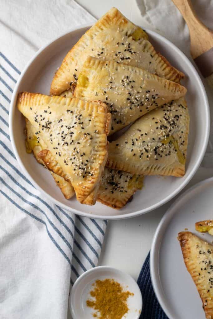 A white plate is piled with baked turnovers with a sesame seed topping.