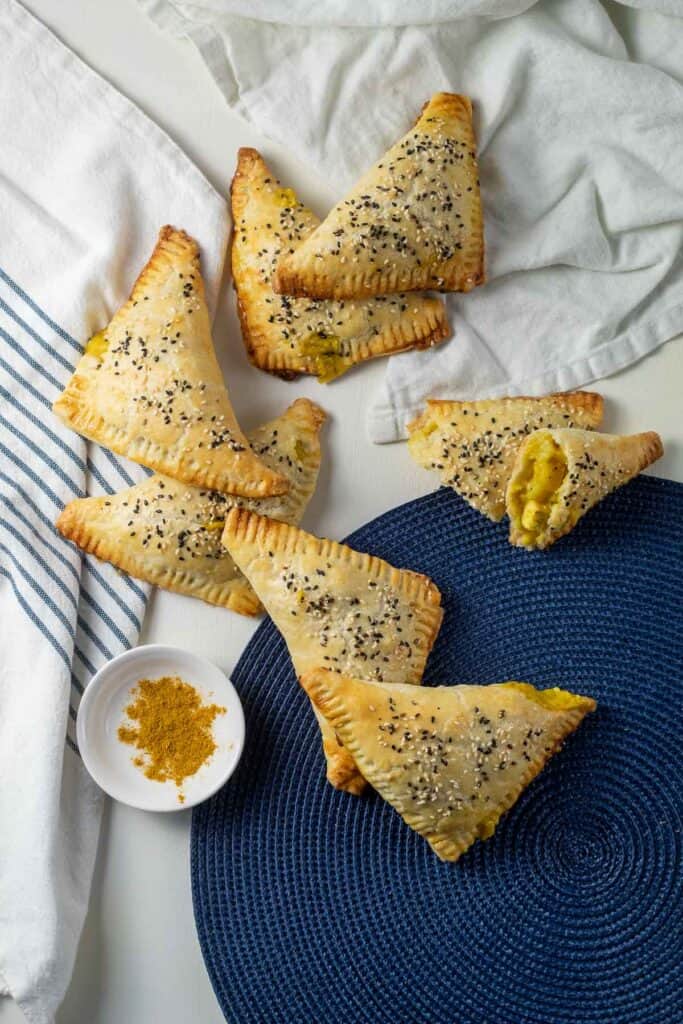 Golden brown turnovers arranged on a blue and white surface alongside a small bowl of curry powder.