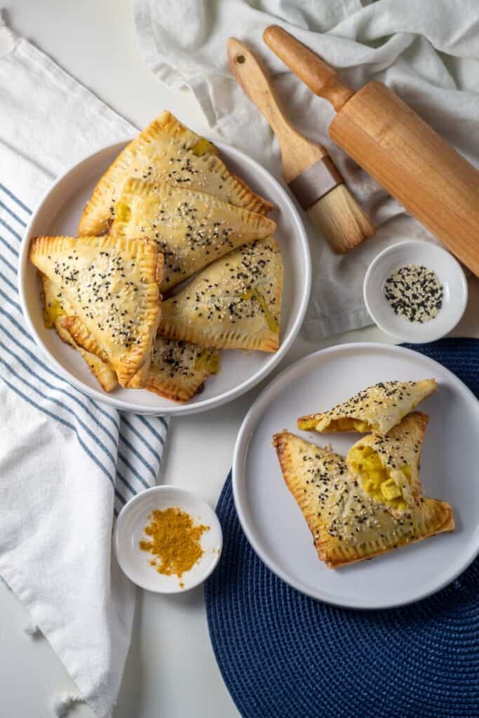 A white plate with a pile of golden turnovers sprinkled with sesame seeds sits alongside a pastry brush, rolling pin, and small bowls of ingredients.