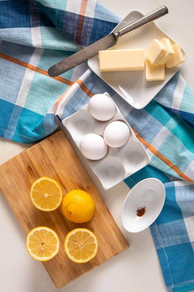 Ingredients for Mom's Microwave Hollandaise, including butter, fresh lemon, and eggs, are arranged on a white surface with a blue plaid towel.