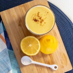 A bowl of yellow sauce sprinkled with orange spice sits on a cutting board next to a sliced lemon half and a small white spoon.