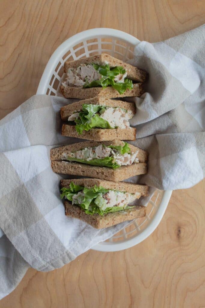 Triangle quarters of a chicken pecan salad sandwich with green lettuce placed in a white plastic basket with a checkered cloth.