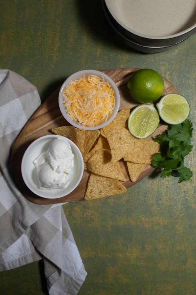 The garnishes for green chile chicken soup with beans, including sour cream, shredded cheese, tortilla chips, limes, and cilantro, arranged on a wood platter.