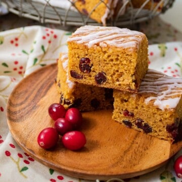 A wooden plate holding three squares of a golden cake studded with cranberries and drizzled with a white glaze.