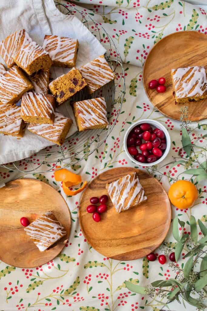 A platter of snacking cake squares alongside three plates with cake, fresh cranberries, and orange peel as garnish.