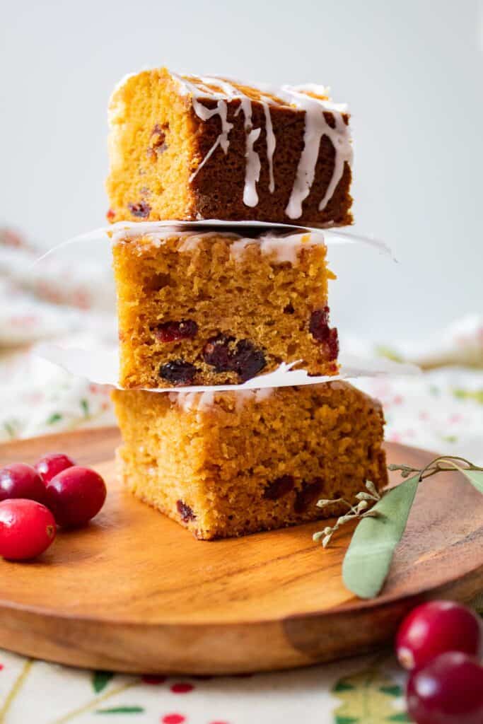 Three squares of a golden cake studded with cranberries and drizzled with a white glaze stacked in a tower on a wooden plate.