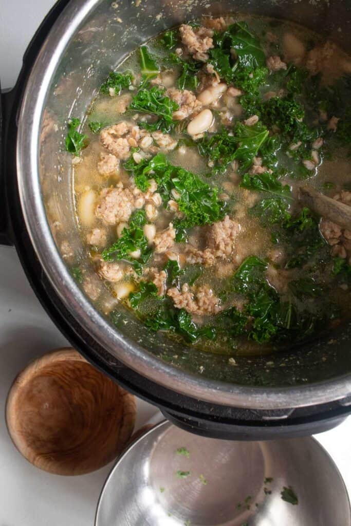 A top-down view of a soup in an instant pot insert with kale, farro, sausage, and white beans visible.