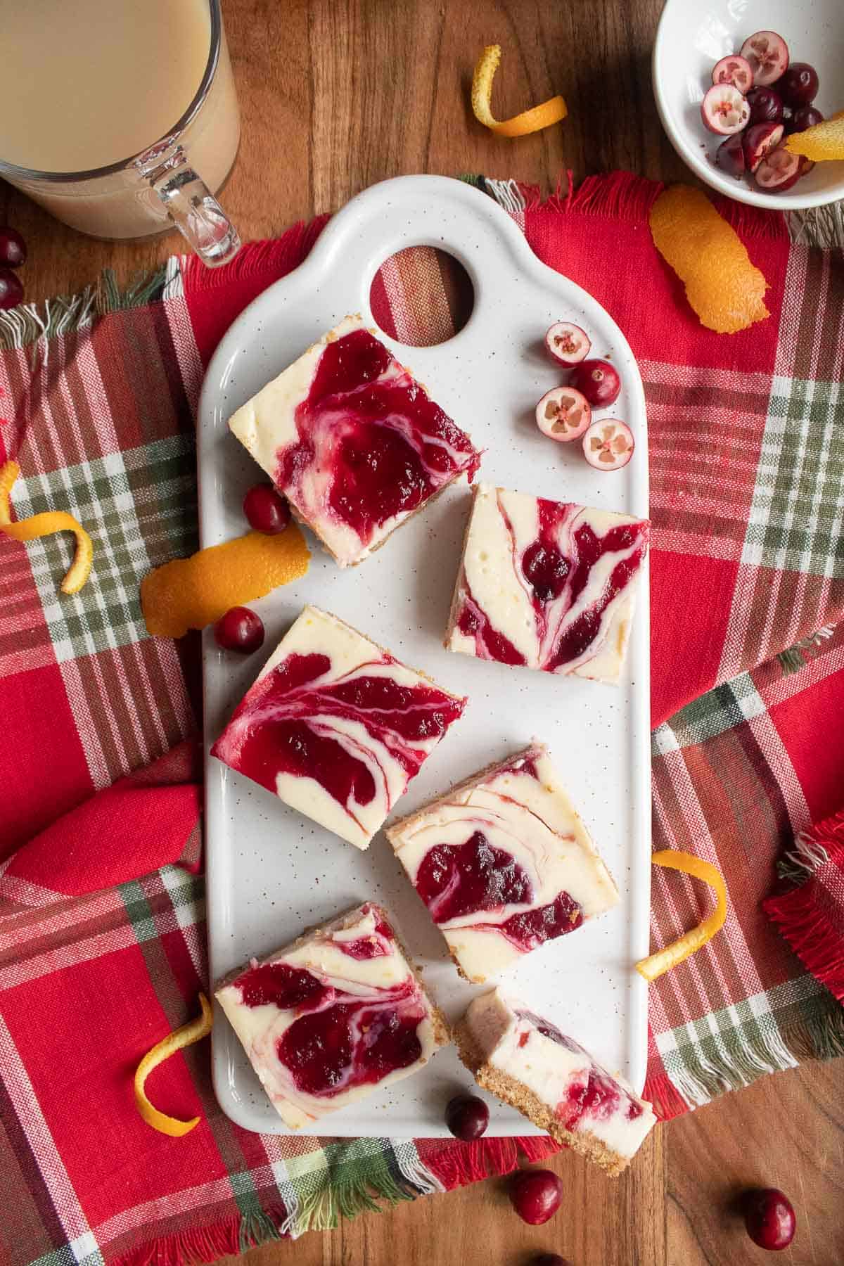 Marbled cranberry cheesecake bars on a white platter for serving surrounded by orange zest, cranberries, and a cup of milky tea.