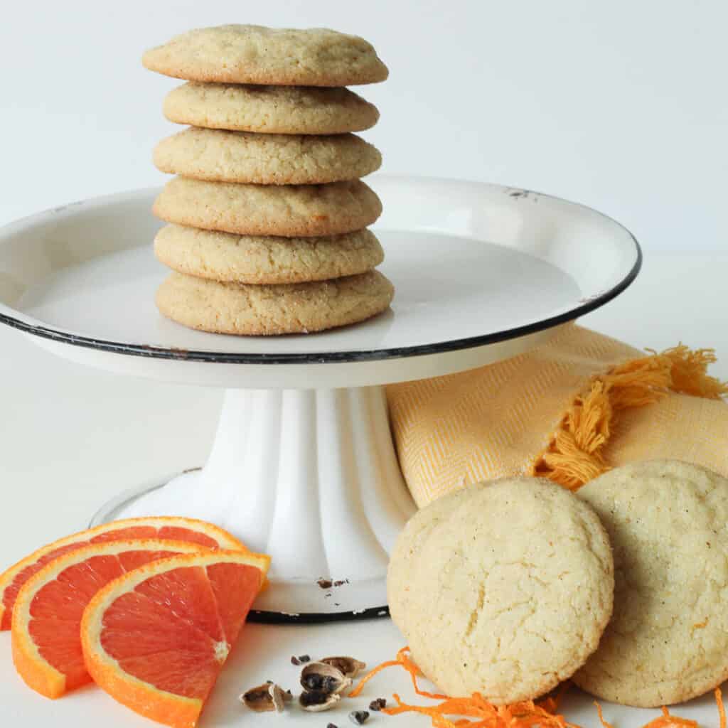 A tall stack of golden edged cookies sitting on a white plate with a black rim. Below are orange slices, cardamom pods, and orange zest.