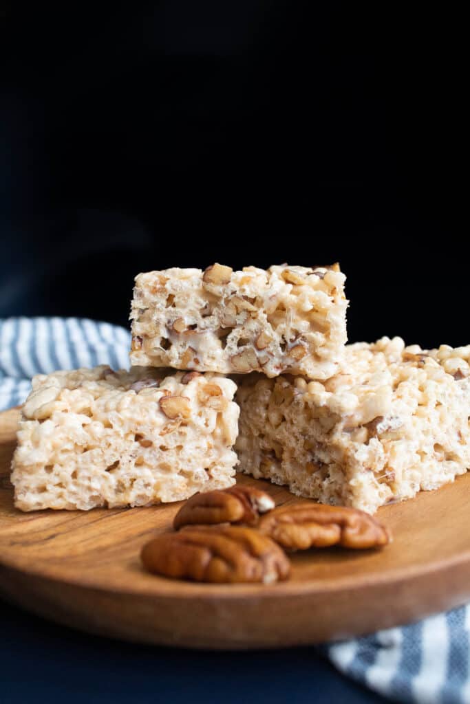 Cut buttered pecan rice krispie treats stacked on a brown plate alongside some pecan halves.
