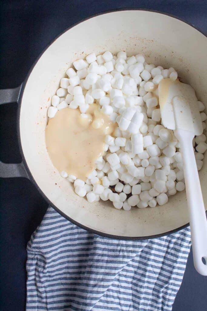 Mini marshmallows and sweetened condensed milk being stirred into the butter and pecan mixture in a large pot.